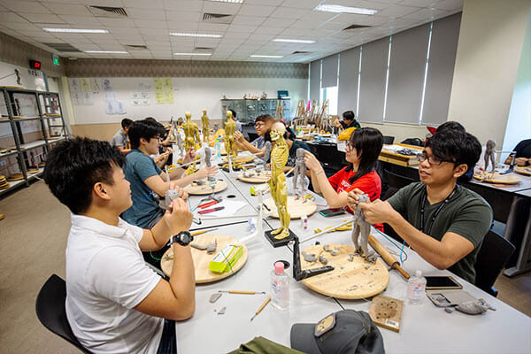 Students at a long table model human figures with gray clay based on miniature skeleton models standing between them.
