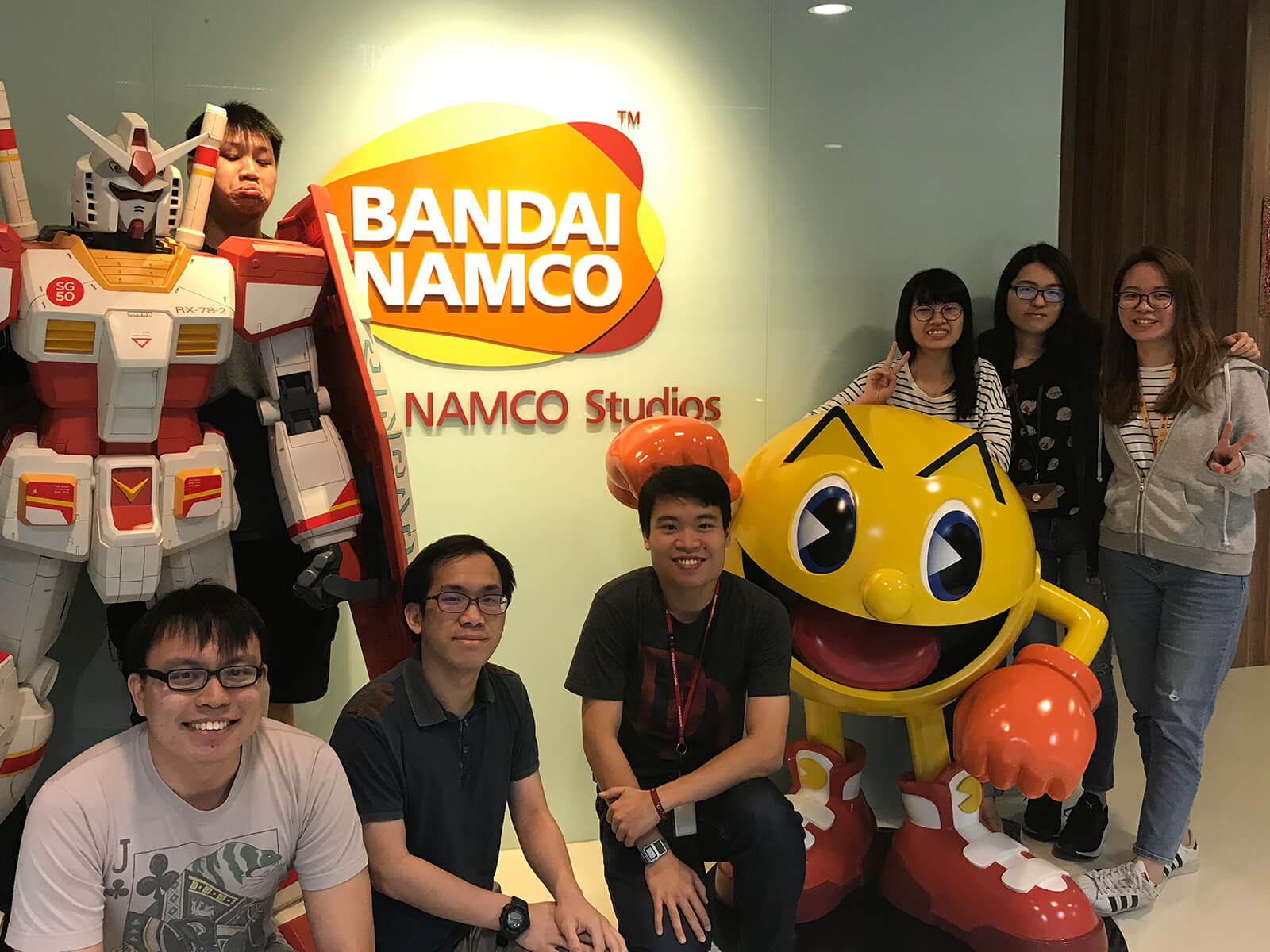 Group photo of eight DigiPen alumni in the lobby of Bandai Namco studio, posing by statues of Pac-Man and a mecha robot.