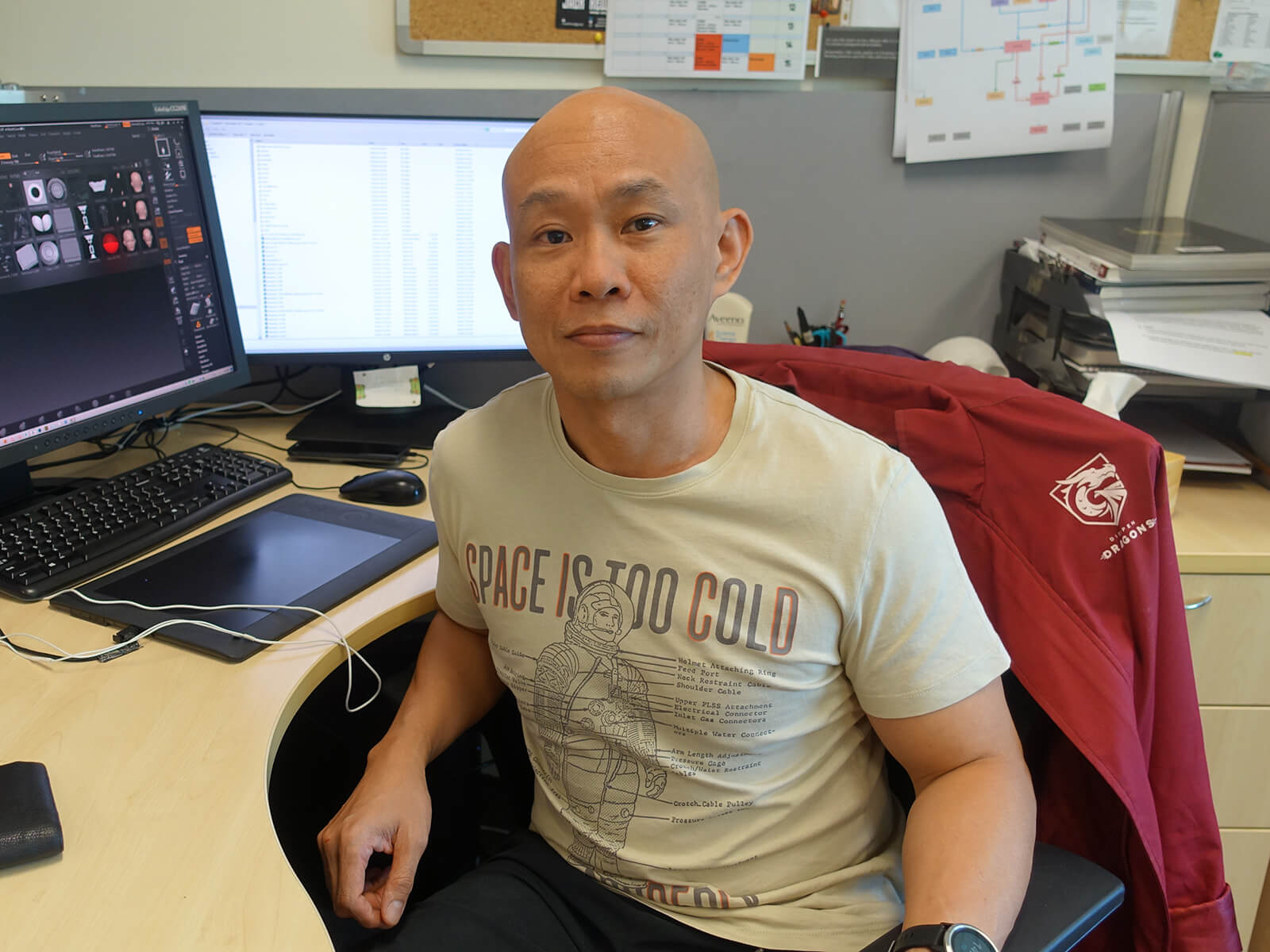 Professor Marc Tan, wearing a tan T-shirt, faces the camera while sitting at his office desk; computer monitor shows thumbnails of his 3D artwork.