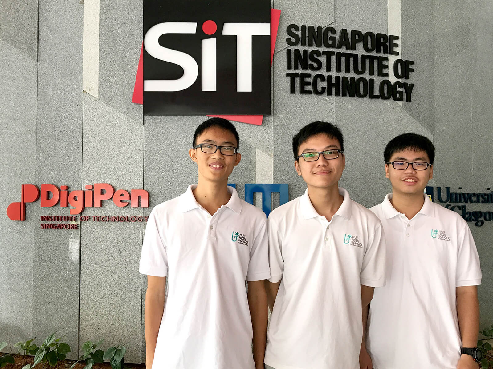 Three students from NUS High School pose for a photo in front of a gray marble wall with the SIT and DigiPen logos