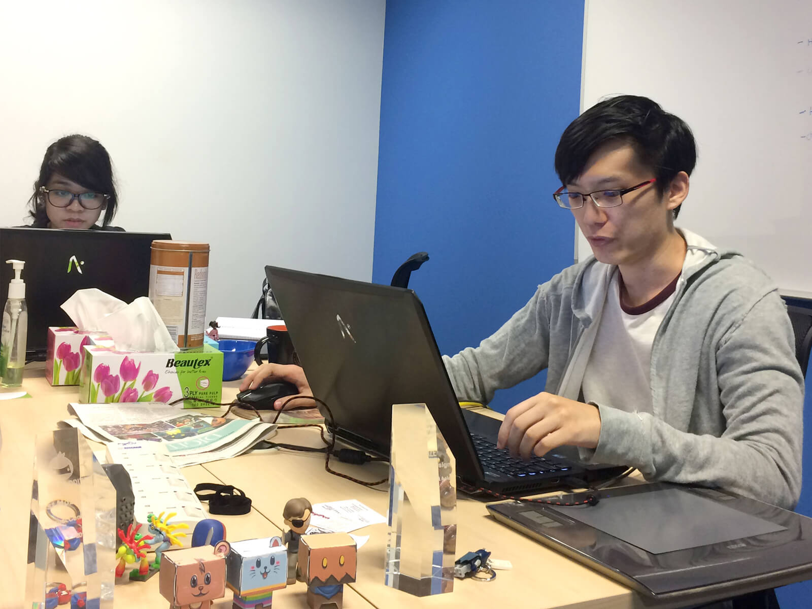 Alumnus Desmond Wong sits at a desk with a laptop computer surrounded by trinkets and awards