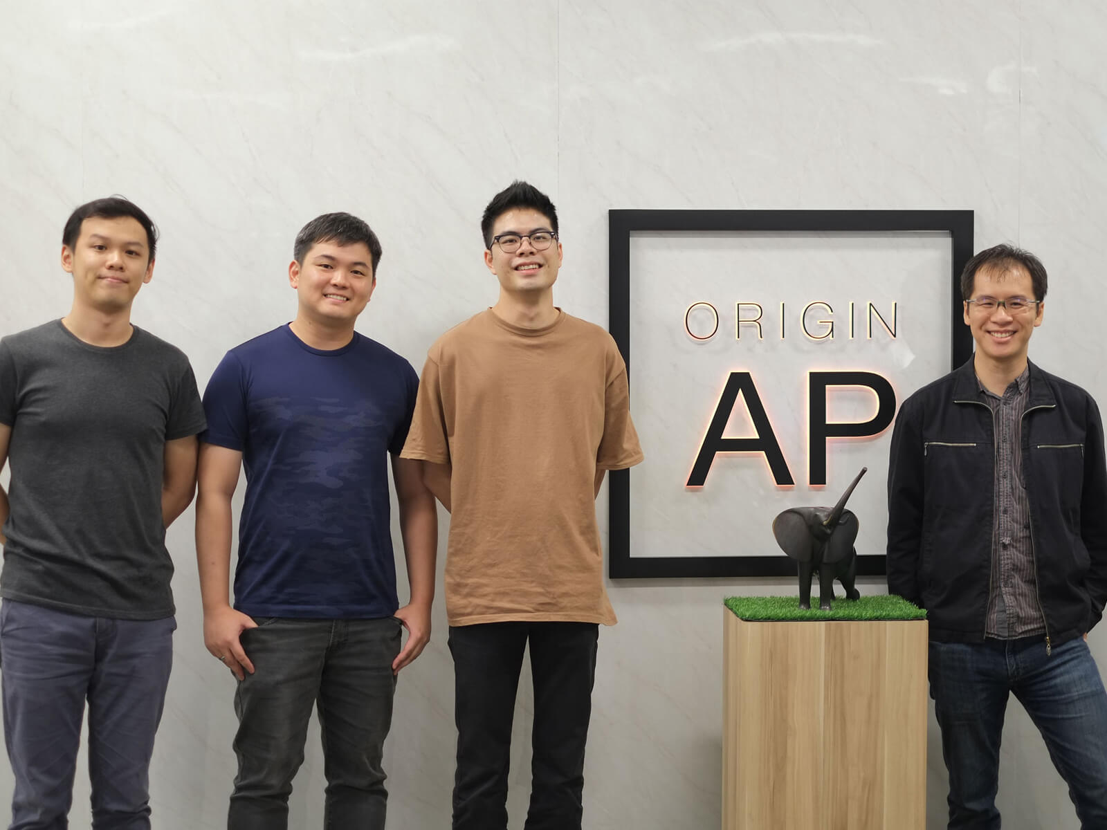DigiPen alumni stand against a white marble wall with the Origin A P logo between them