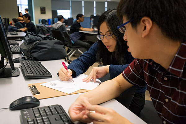 A student writes on a piece of paper at a desk with computers on it as another student looks on.