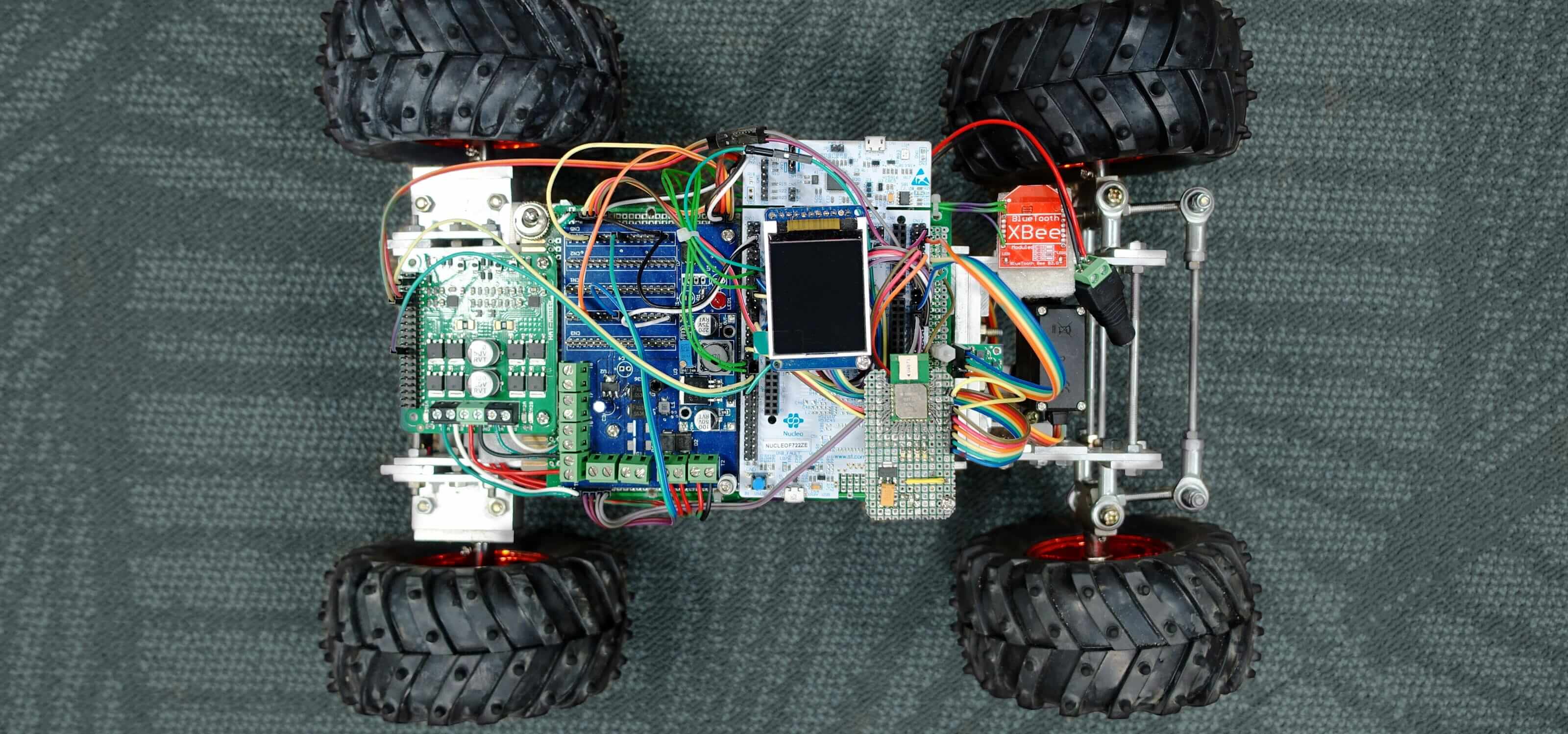 A model racing car seen from above with textured black tires and colorful exposed wiring and circuit board.