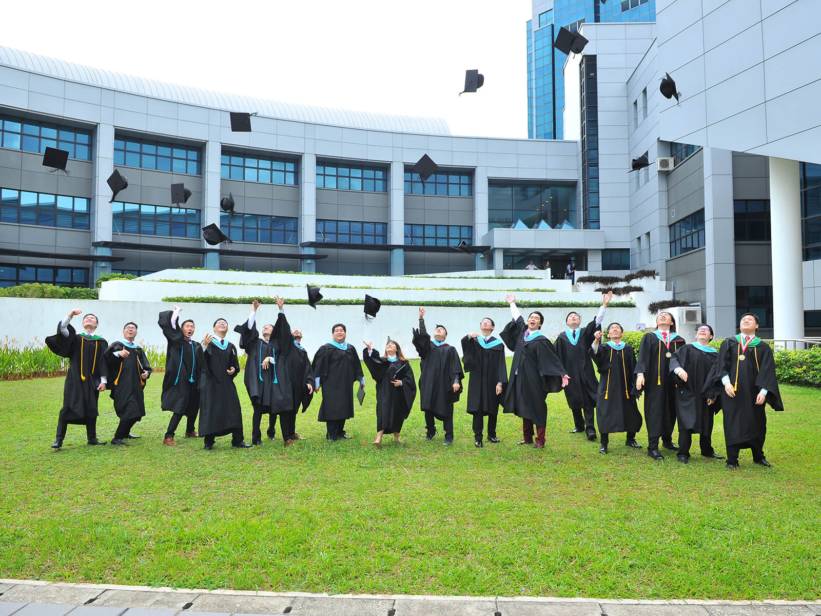 Several lined-up graduates throw their graduation caps into the air while standing outside on the grass.