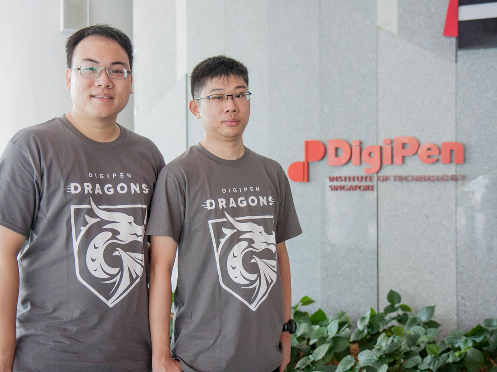 Graduates Tan Tien Wei Keith and Yap Teng Hong pose for a photo in gray DigiPen Dragons t-shirts next to the DigiPen building